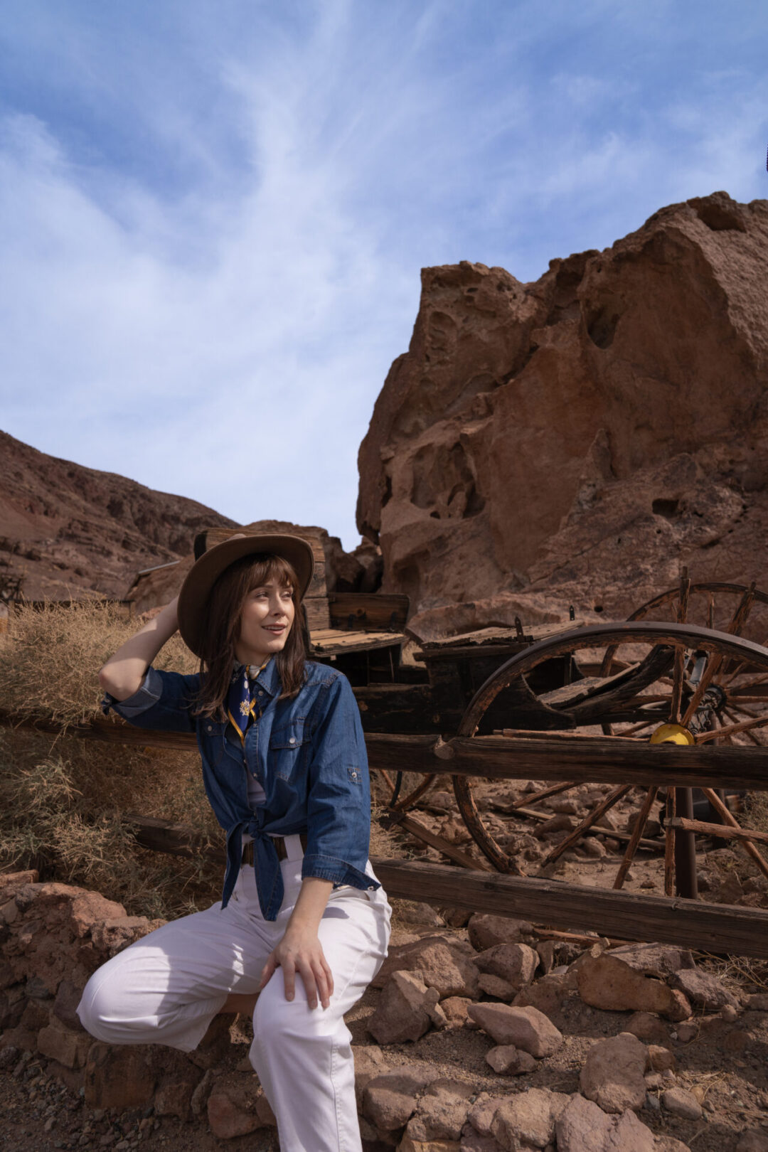 Travel Blogger Jordan Gassner holding her western hat while squatting in front of a vintage wooden wagon at Calico Ghost Town in California