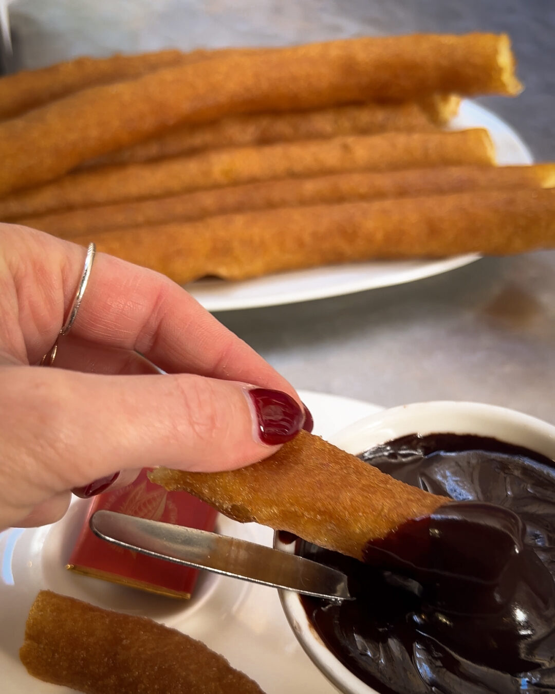 A woman's hand with burgundy nail polish dipping a piece of a churro in dark chocolate from Chocolatería Valor in Madrid, Spain