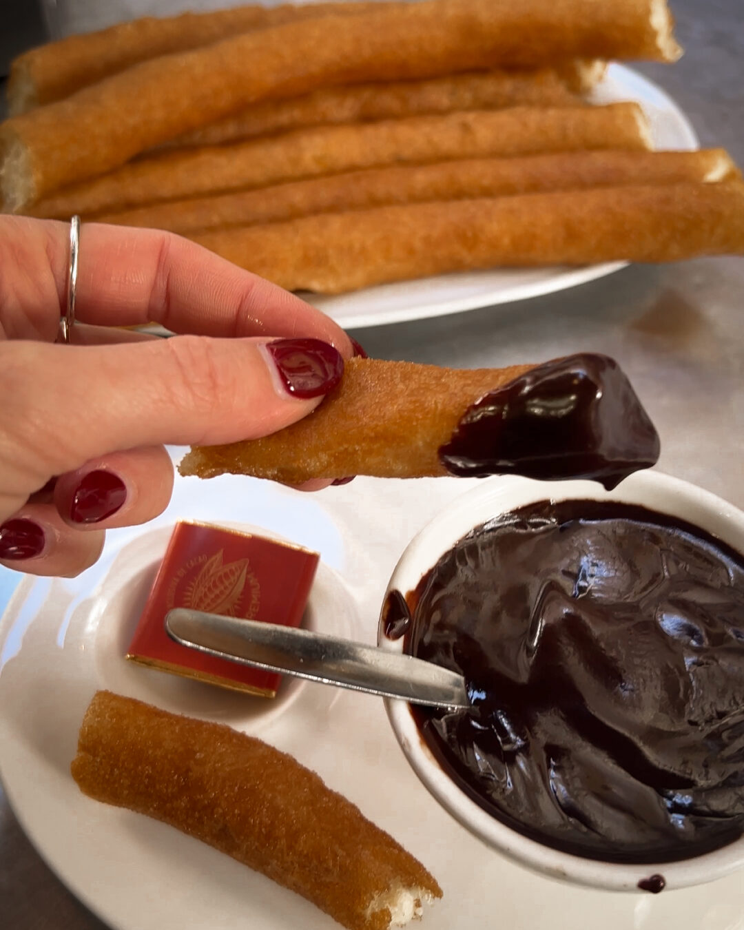 A woman's hand with burgundy nail polish holding a piece of a churro dipped in dark chocolate from Chocolatería Valor in Madrid, Spain