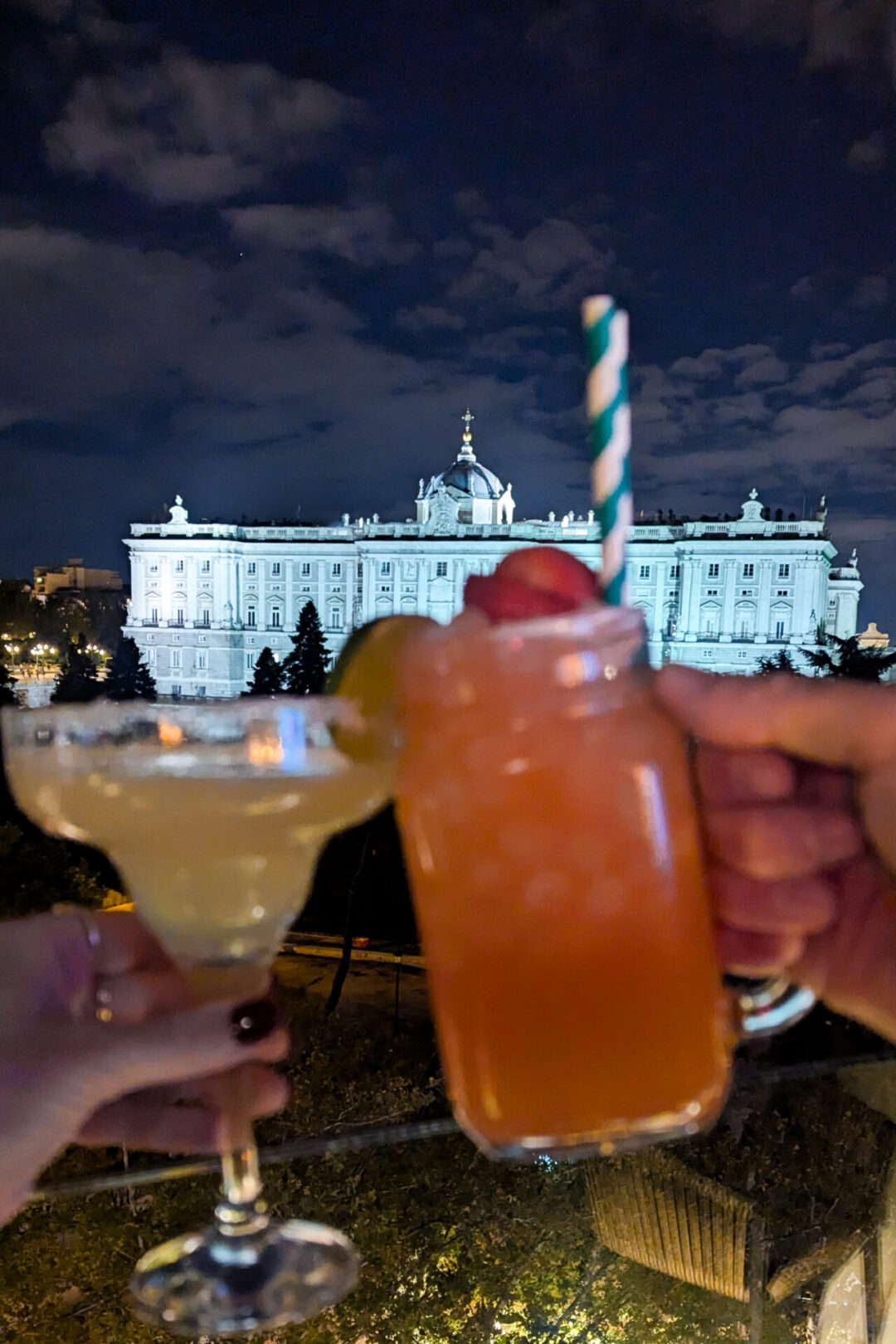 A couple cheers'ing two cocktail glasses with a view of the Royal Palace of Madrid behind them