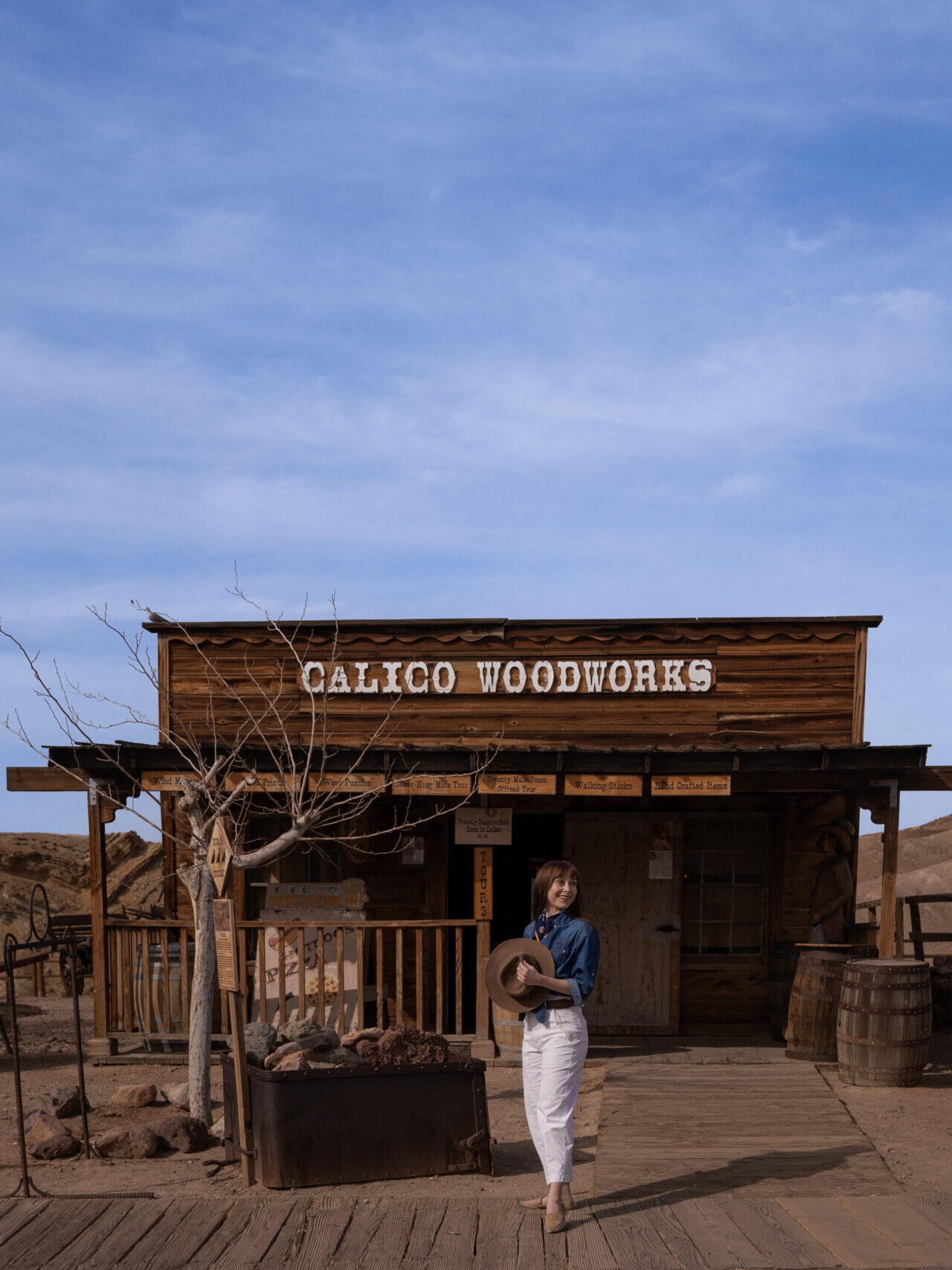 Travel Blogger Jordan Gassner holding her western hat and looking over her shoulder and smiling in front of Calico Woodworks store at Calico Ghost Town in California