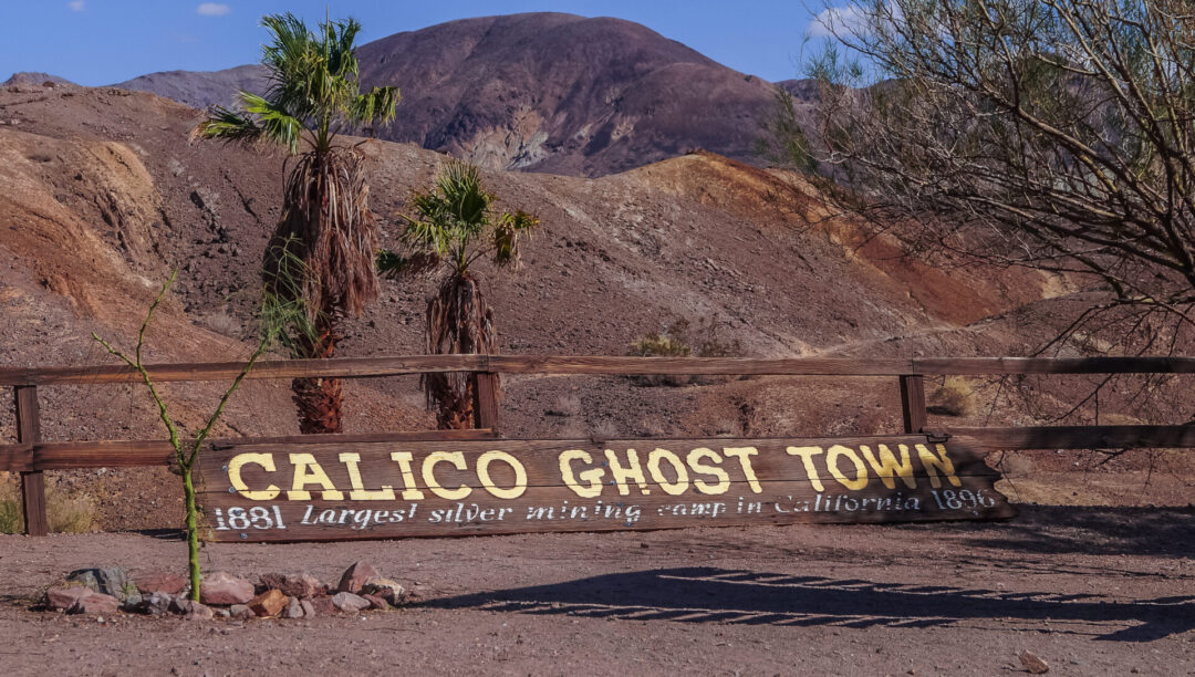 A sign reading "Calico Ghost Town: 1881 Largest silver mining town in California 1896" laying on a path in the desert with colorful mountain ranges just behind
