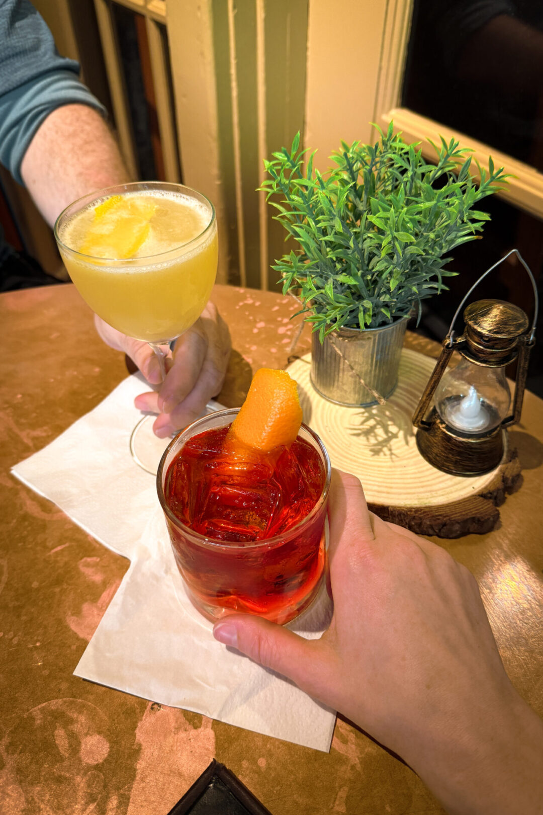 A woman and man's hands holding a lemon cocktail and a negroni from The Ahwahnee Hotel Bar