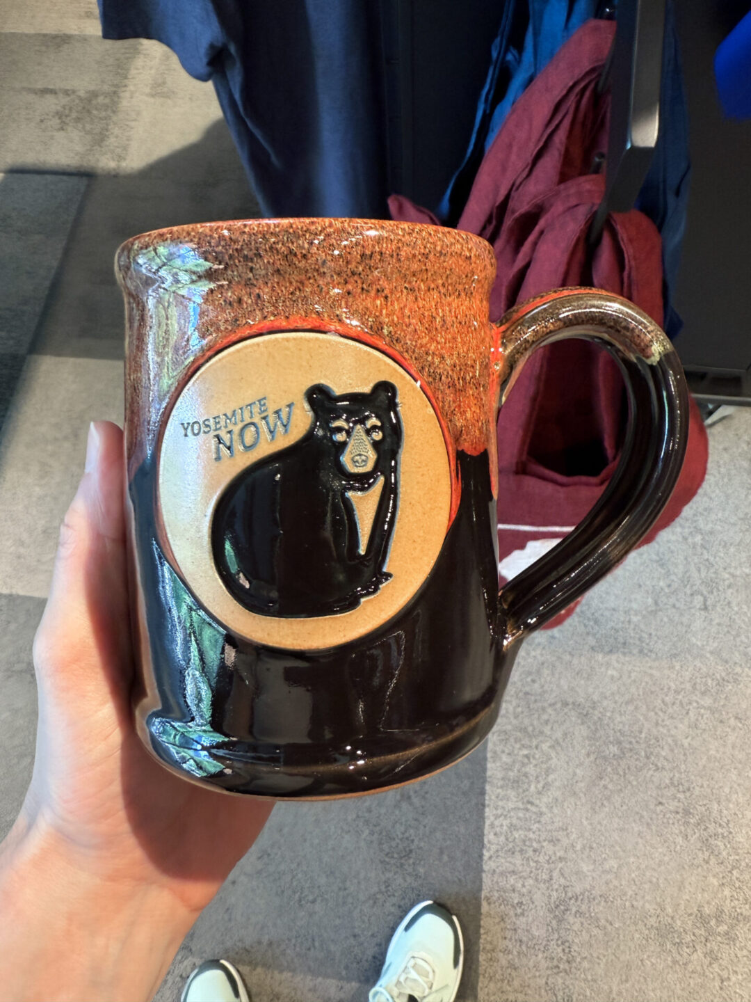 A woman's hand holding up a mug with "Yosemite Now" and a bear on it inside the Yosemite National Park Welcome Center