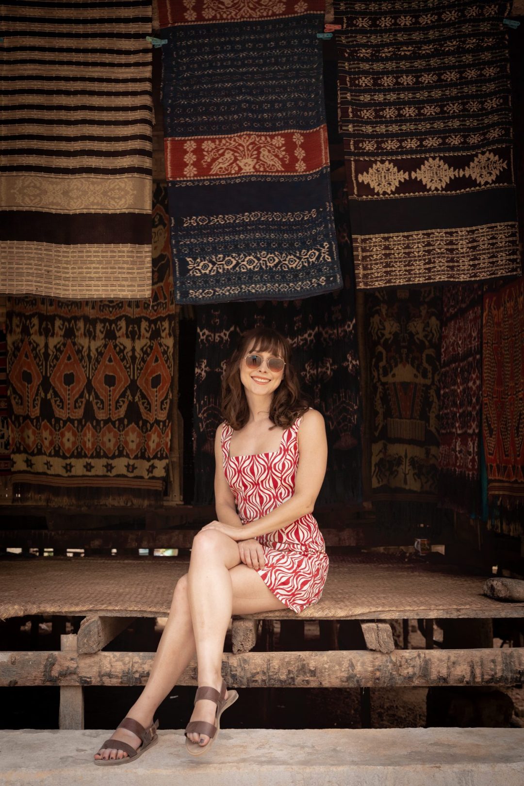 Travel Blogger Jordan Gassner sitting on a mat inside a home surrounded by lots of hanging ikat textiles in Prailiu Village in Sumba, Indonesia
