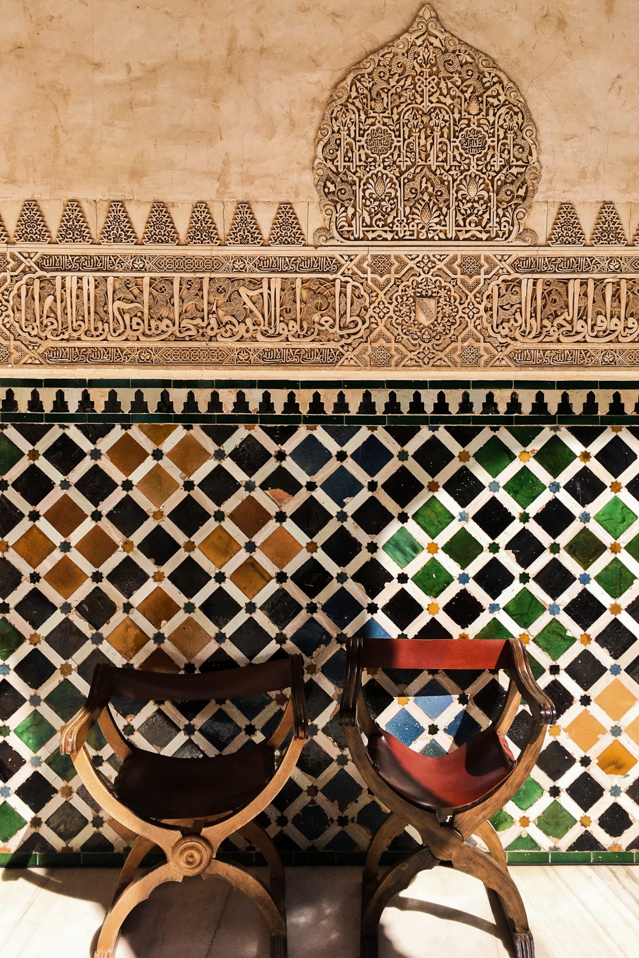 Two wood and fabric chairs sitting against a colorful tile and carved wall in the Moorish design in Andalusia, Spain