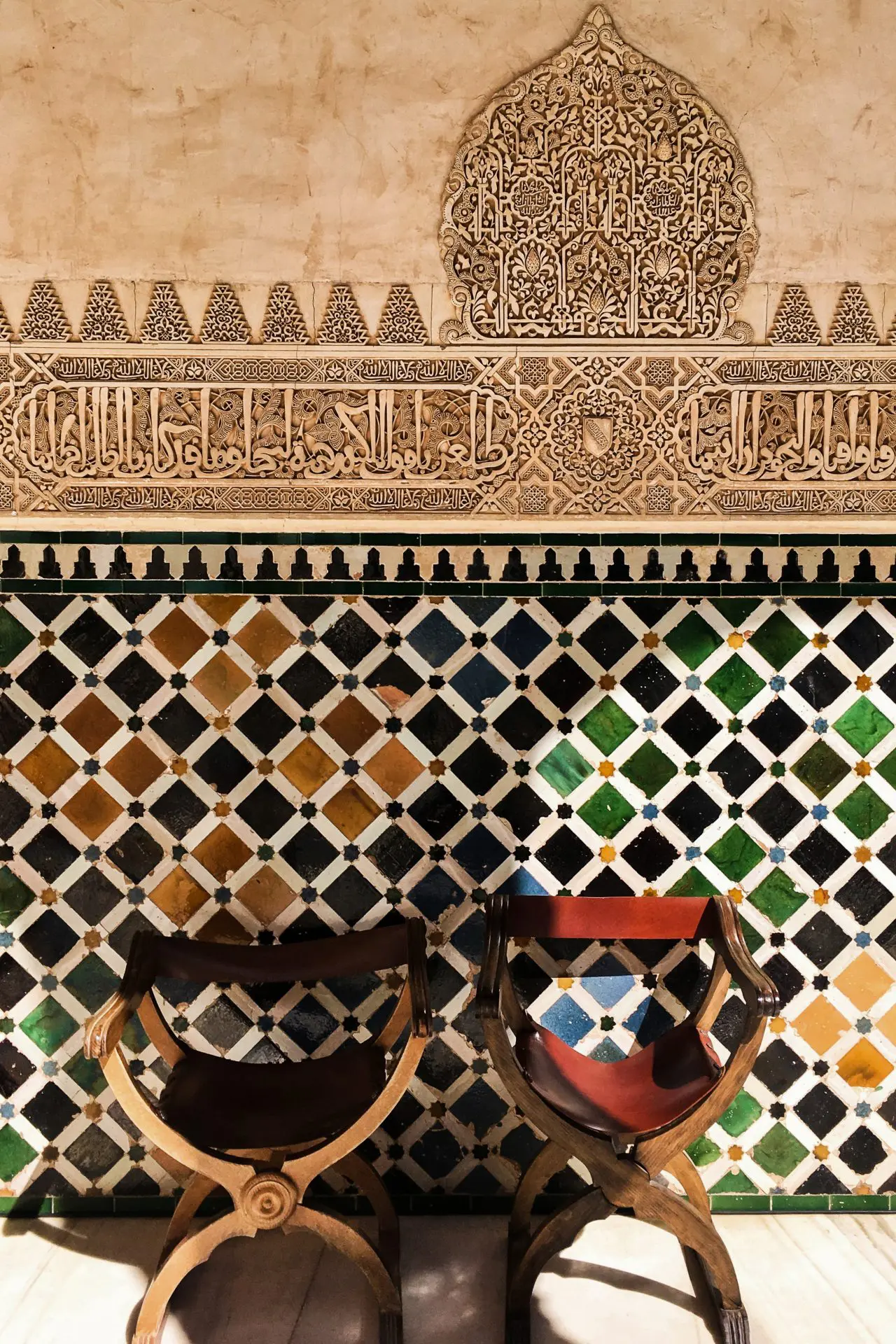 Two wood and fabric chairs sitting against a colorful tile and carved wall in the Moorish design in Andalusia, Spain
