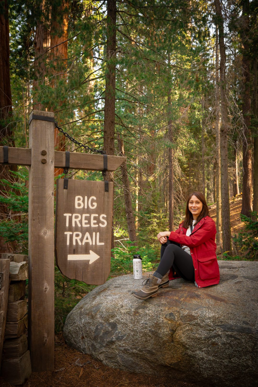 Travel Blogger Jordan Gassner smiling from the top of a rock near the "Big Trees Trail" signpost inside Sequoia National Park