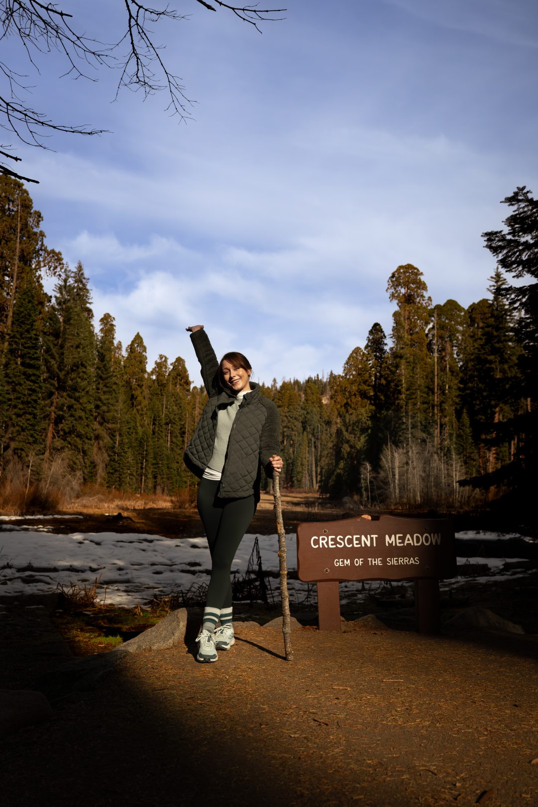Travel Blogger Jordan Gassner smiling with one hand in the air while holding a walking stick in front of Crescent Meadow during a Sequoia National Park Trip