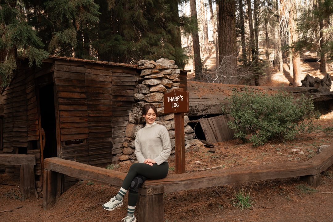 Travel Blogger Jordan Gassner smiling while sitting on a bench in front of Tharp's Log in Sequoia National Park Trip