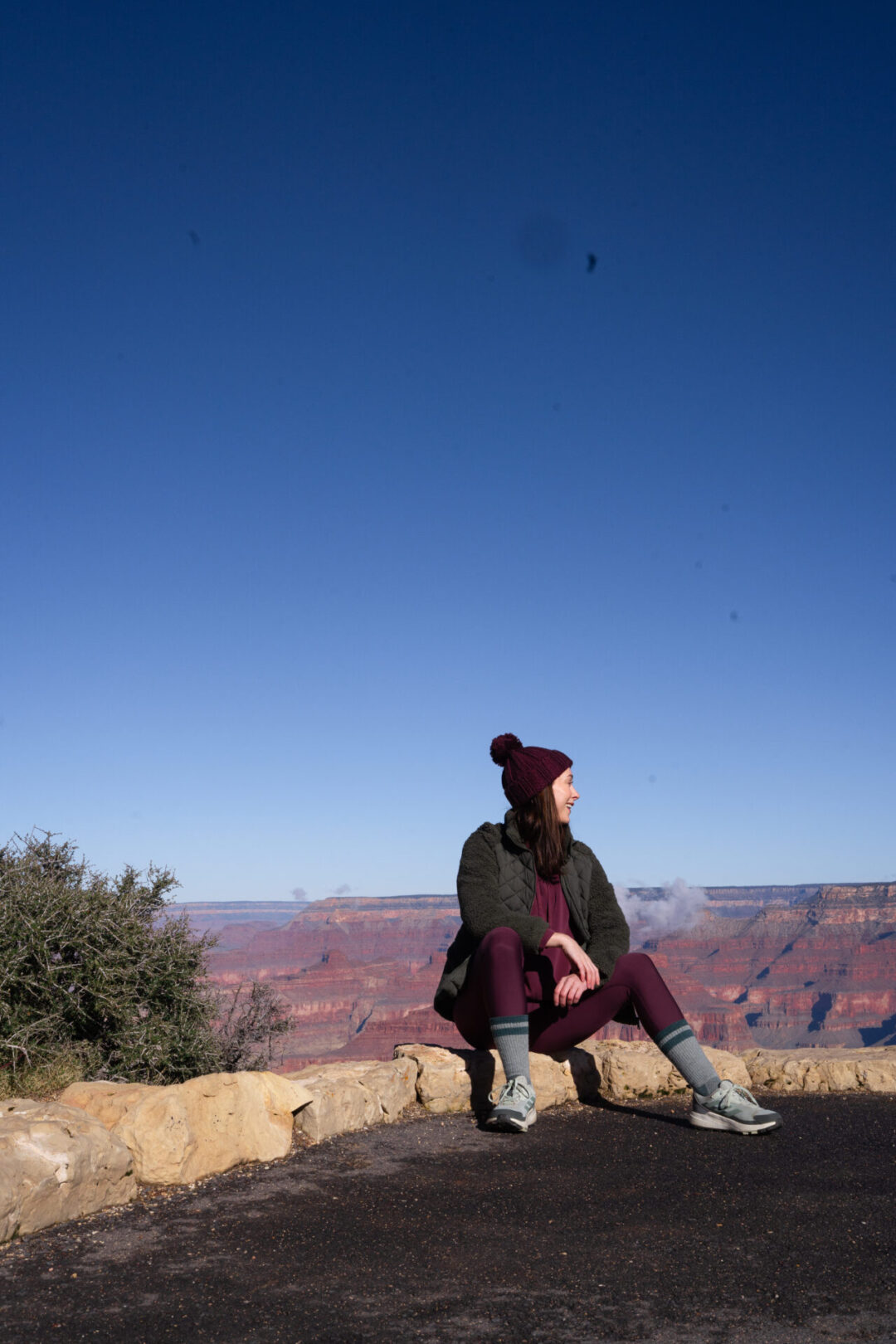 Travel Blogger Jordan Gassner wearing a winter jacket and a cranberry athleisure set while sitting on a rock at Maricopa Point in the Grand Canyon in Arizona, USA