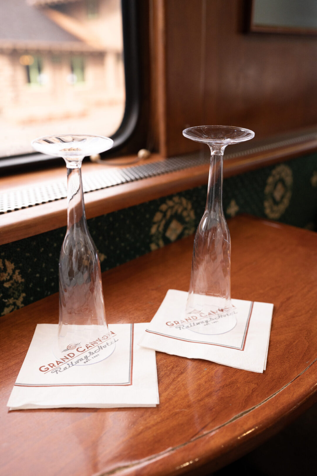 Two plastic champagne flutes and "Grand Canyon Railway & Hotel" branded napkins sitting on a luxury wood tray table inside a train car