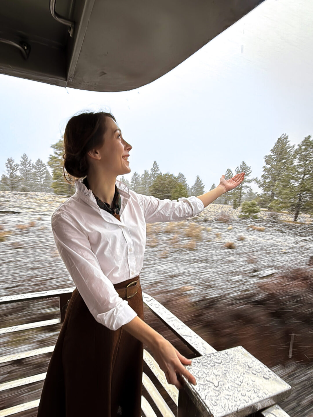 Travel Blogger Jordan Gassner smiling while holding her hand out to catch snow while standing on the back observation deck aboard the Grand Canyon Railway while in motion