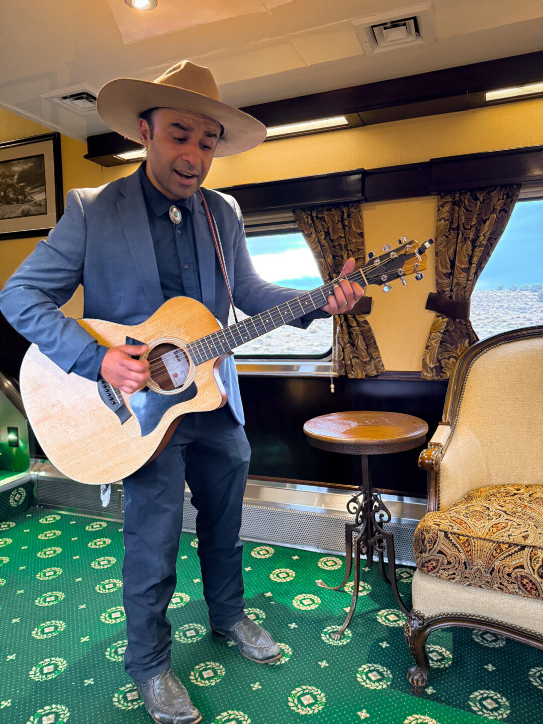 A musician in a blue suit singing and playing guitar inside a luxury parlor car aboard the Grand Canyon Railway