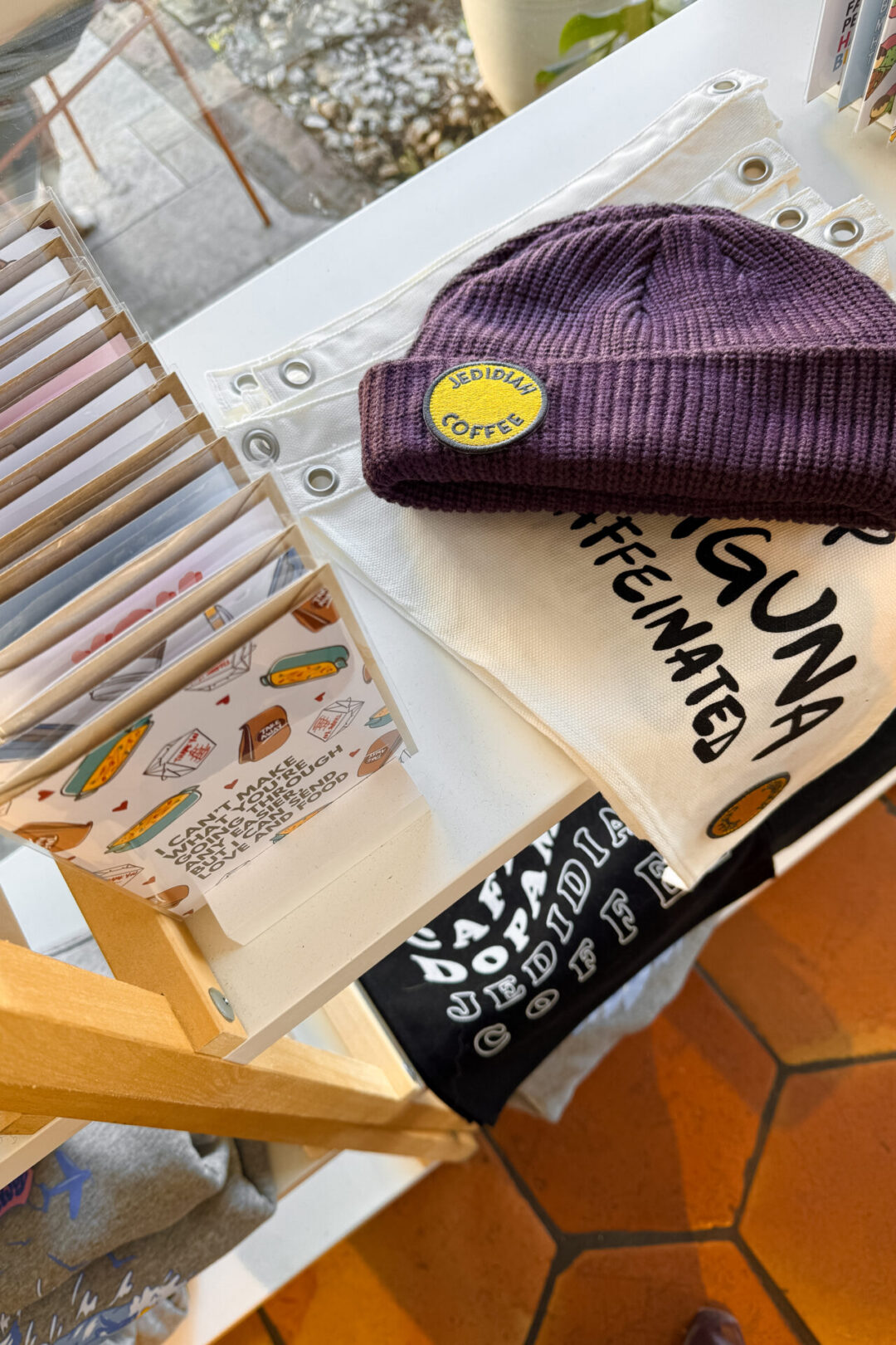 A purple beanie with a yellow "Jedidiah Coffee" logo overlaying more coffee merch in Laguna Beach, California