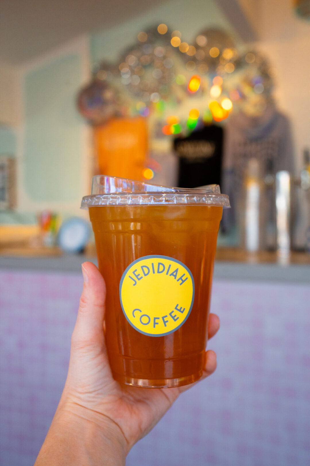 Travel Blogger Jordan Gassner holding a "Jedidiah Coffee" branded iced tea in front of a light purple coffee counter Laguna Beach, California
