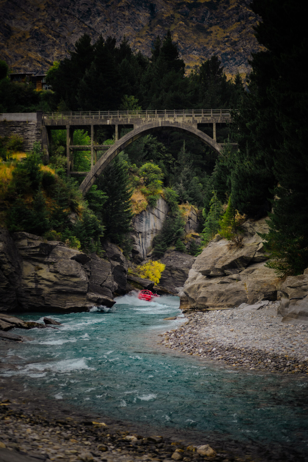A red jet boat going gliding through the narrow slot canyons of the Shotover River underneath a bridge in Queenstown, New Zealand